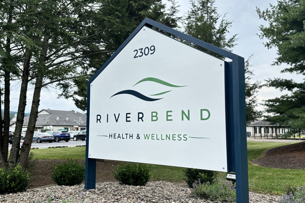 A white sign reads 2309 RIVERBEND HEALTH &amp; WELLNESS with green and blue wave graphics, standing among rocks, plants, and trees under a cloudy sky.