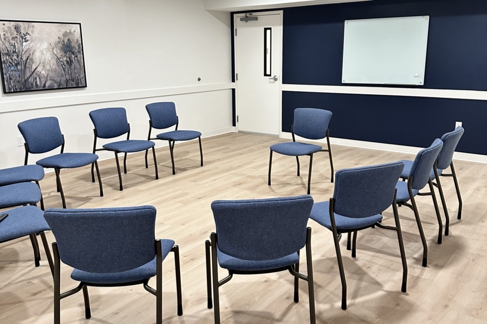 A circle of blue chairs is arranged in an empty room with light wood floors, a whiteboard on a dark blue wall, a painting, and overhead lights.
