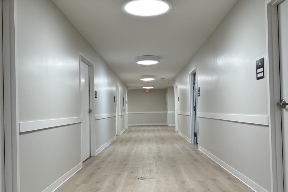 A clean, empty hallway with light wood floors, white walls, and several closed doors on both sides. Circular ceiling lights are evenly spaced, and an exit sign is visible at the far end.