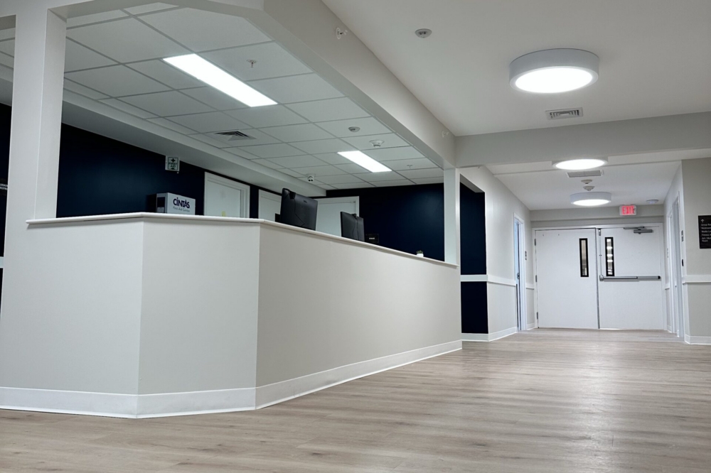 A modern, clean hallway with light wood floors leads to a reception desk with computer monitors. The area has white walls, navy accents, and round ceiling lights, creating a bright and welcoming atmosphere.