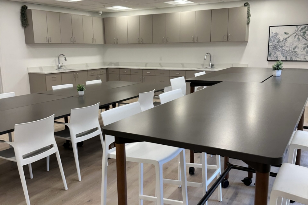 A modern break room with long tables, white chairs, light wood floors, beige cabinets, two sinks, small plants on tables, and a framed artwork on the wall. Bright, evenly lit with ceiling lights.