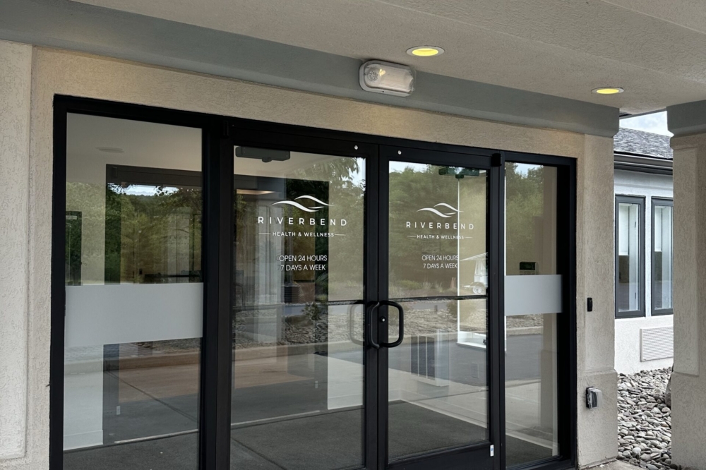 Glass double doors with handles lead into a building labeled Riverbend with a covered entryway. The doors have white text and trim, with a concrete sidewalk and stone accents visible outside.